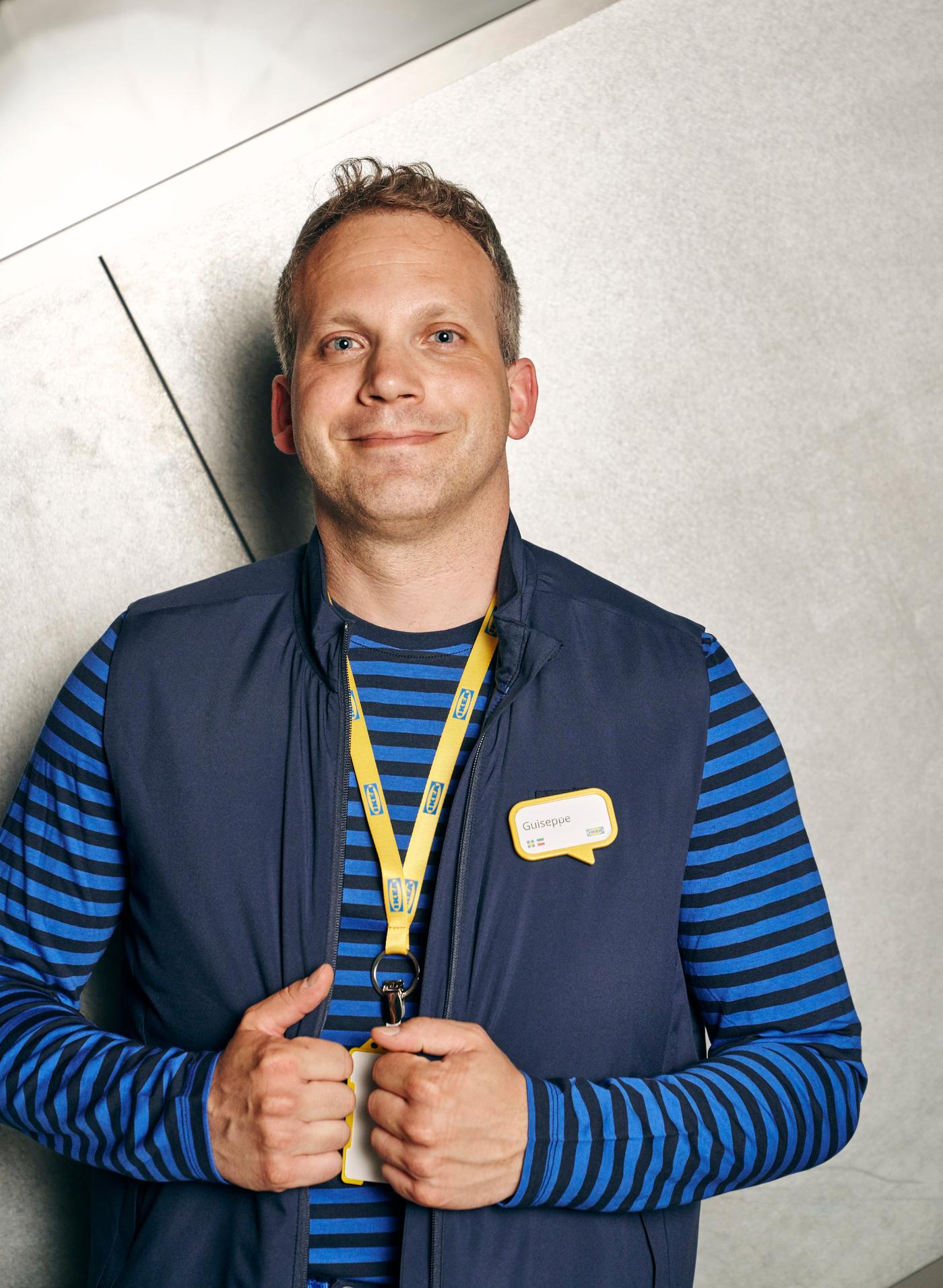 A man smiles as he looks at the camera. He is wearing a striped blue top, a navy gillet and a yellow lanyard.