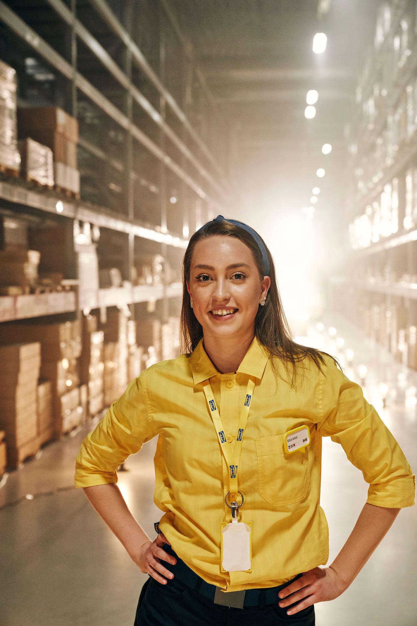 A woman stands in the aisle of an IKEA store. She is wearing a yellow shirt and a lanyard.