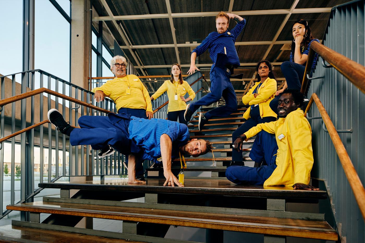 A group of IKEA co-workers stand, set and jump around on a wooden staircase in an IKEA store.