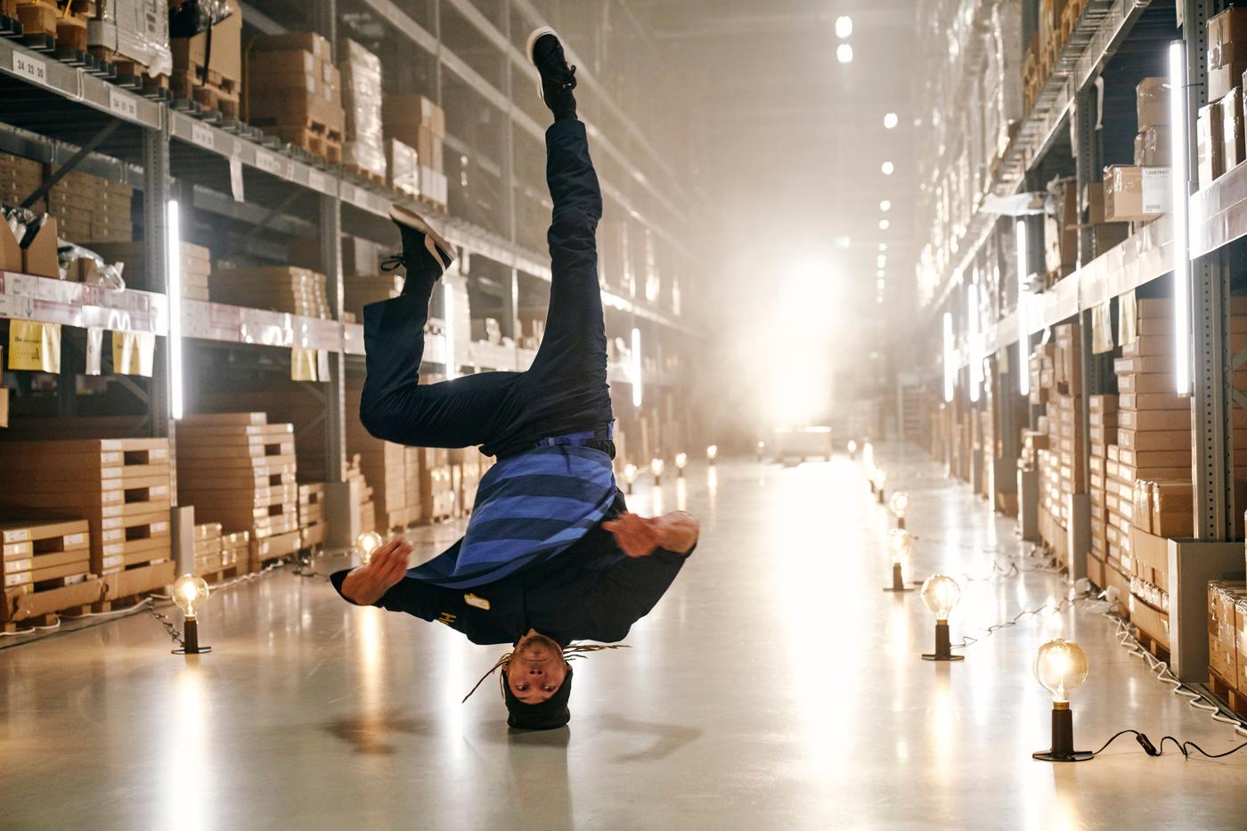 A man stands on his head in the aisle of an IKEA store. There are lamps lit along the aisle.