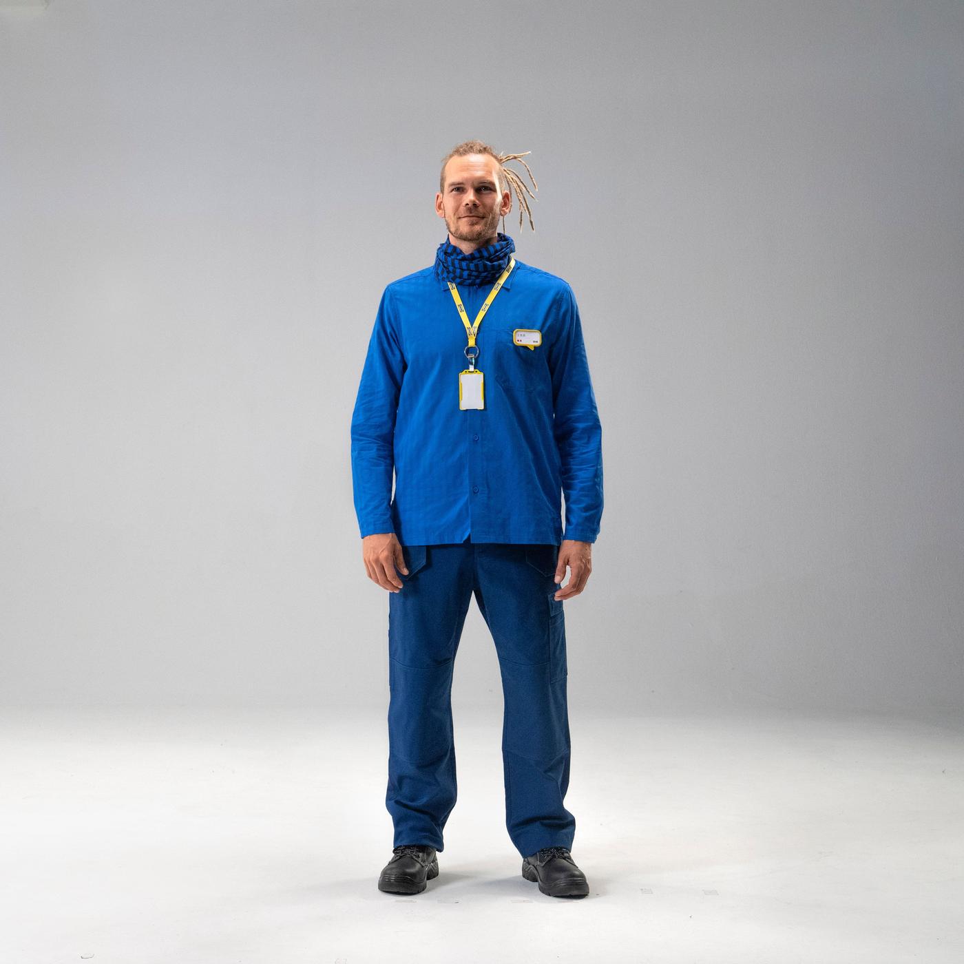 A man stands looking at the camera. He is wearing blue trousers and top with a lanyard around his neck.