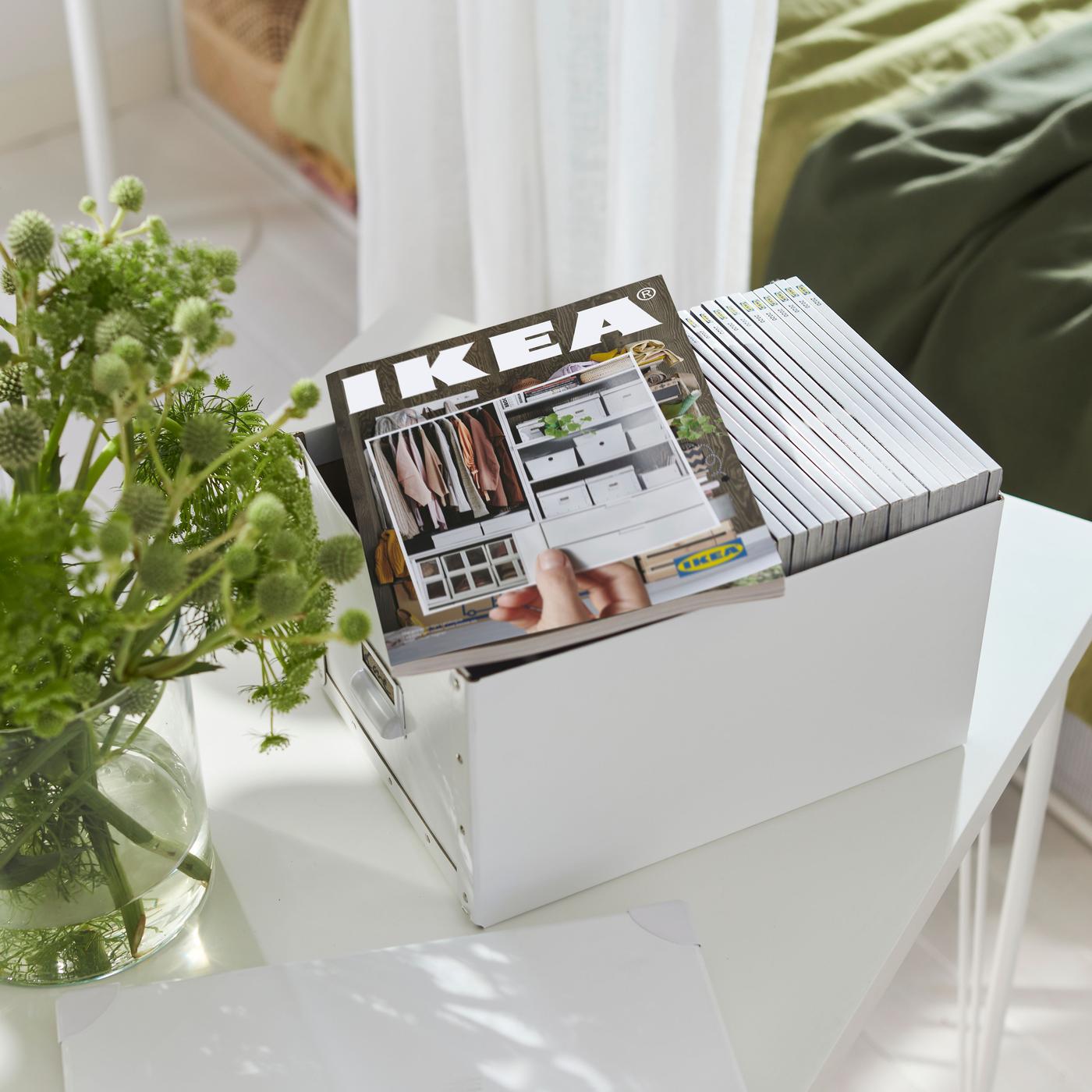 A white box filled with catalogues. There is a catalogue on top. There is also a vase of flowers to the left.