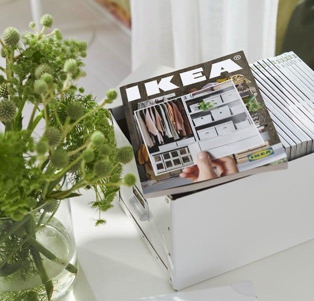 A white box filled with catalogues. There is a catalogue on top. There is also a vase of flowers to the left.
