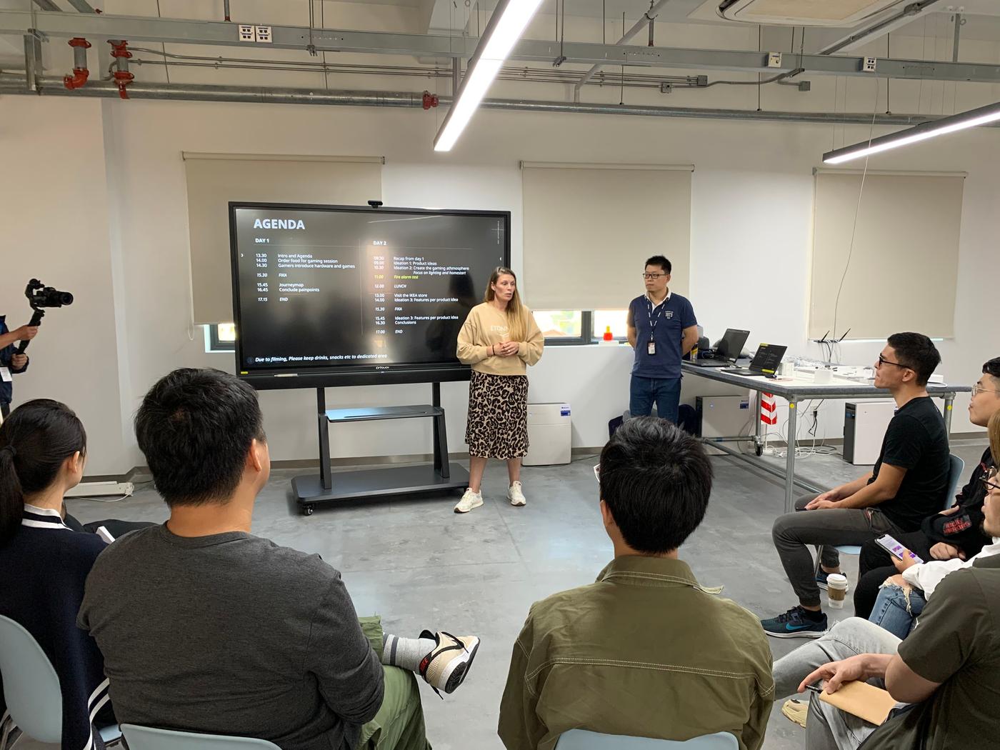 A person stands in a room in front of a large monitor. There is another person standing, while the others all sit and watch.