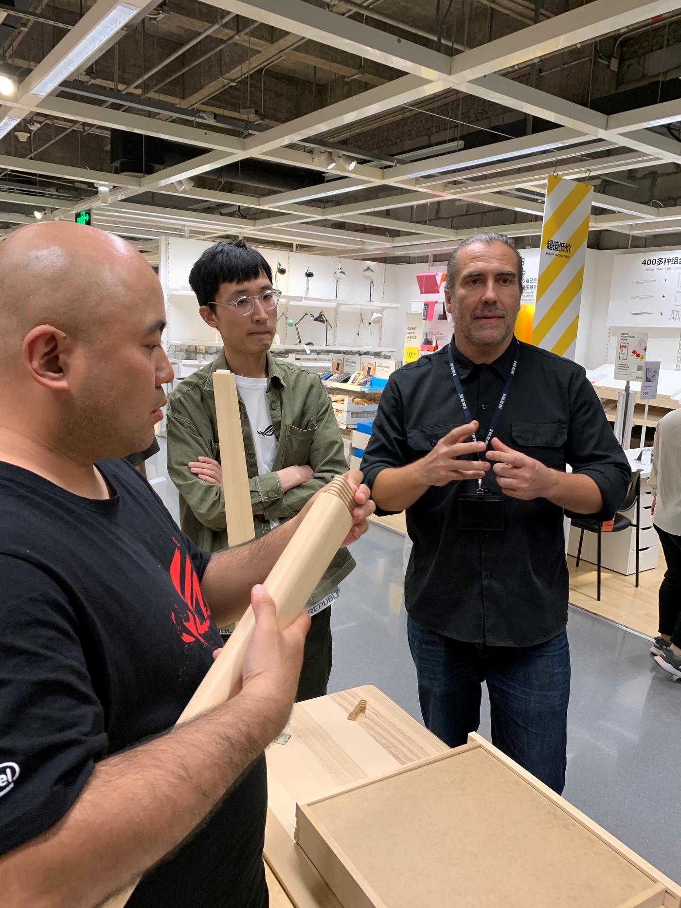 Three people stand in a room. One is talking, while another is looking at a piece of wood.