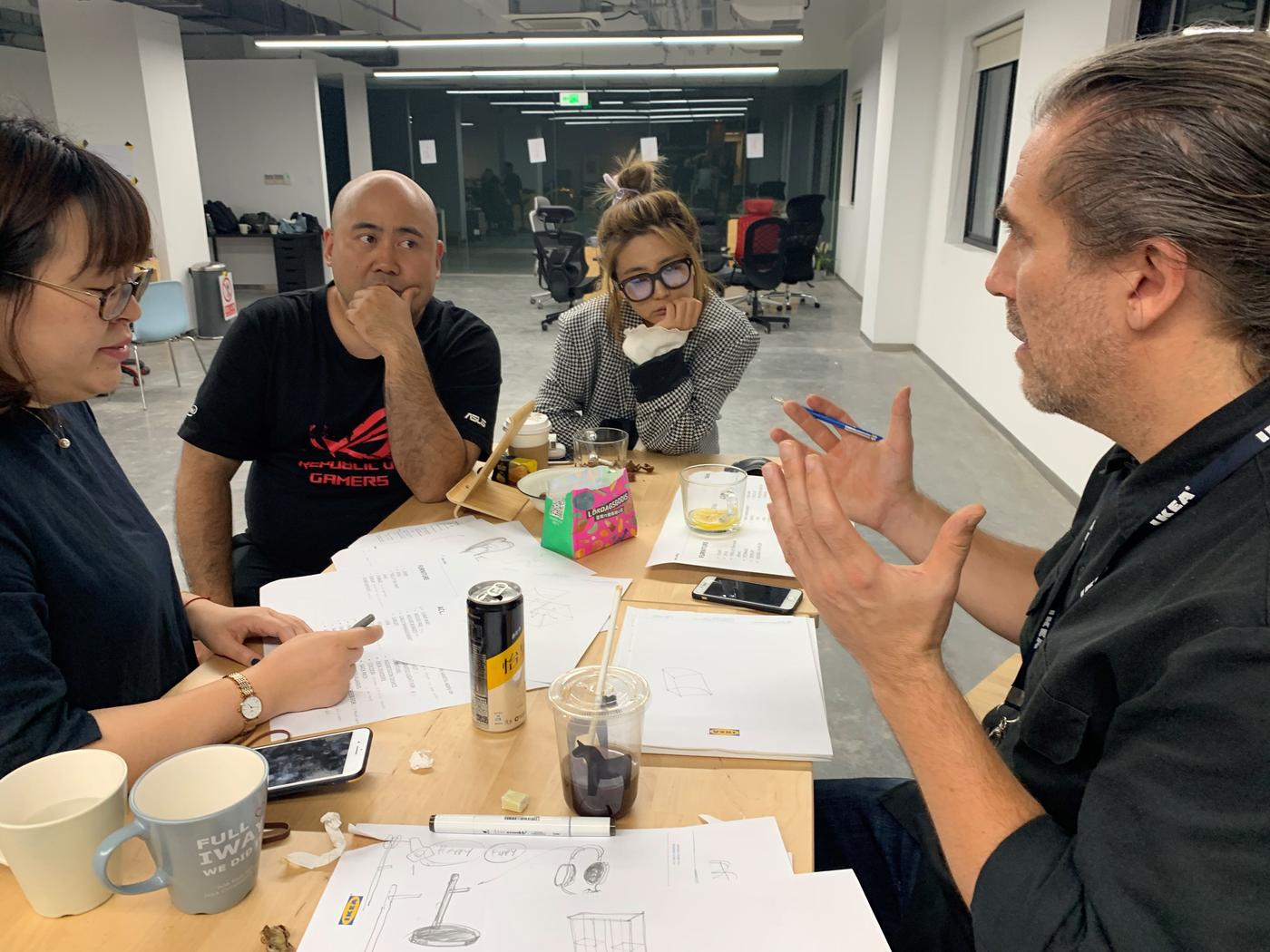 Four people sit around a table. The table is covered with papers and drinks. One person explains while the others listen.