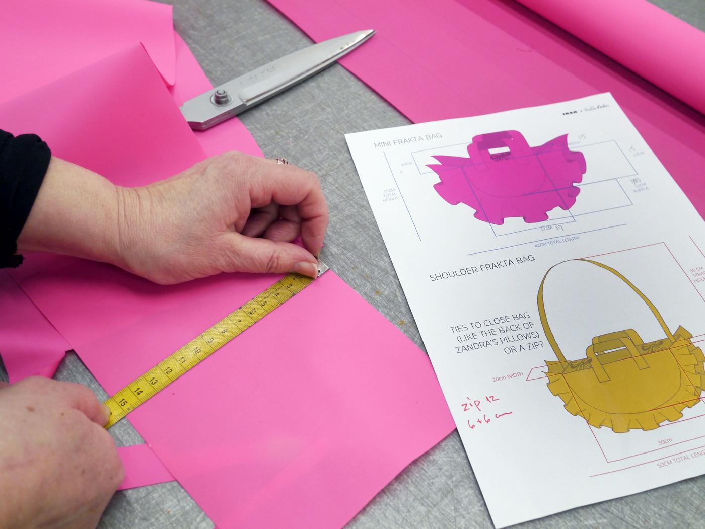 A pair of hands measure a piece of pink fabric, following a bag pattern. There are a pair of scissors too.