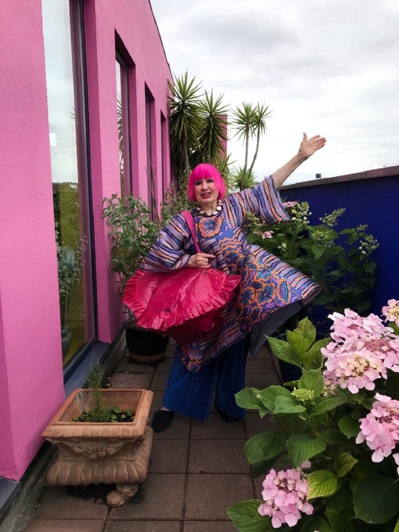 A woman stands on the balcony of a pink building. She has one hand in the air and is smiling. She has a pink bag on her shoulder.