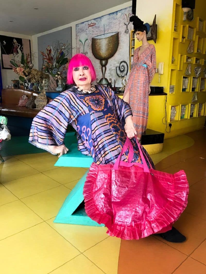 A woman leans on a big green Z shaped chair. She is holding a pink bag. The room has a yellow floor.