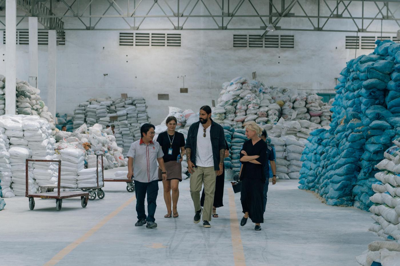 Four people walk through a large warehouse. There are sacks in blue and white stacked around them.