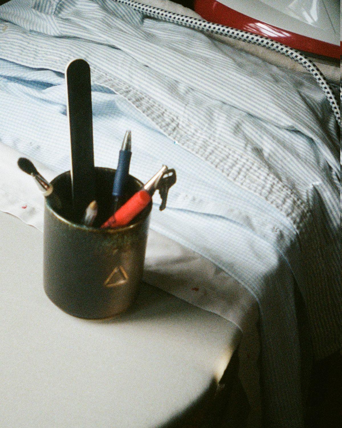 An ironing board with shirts, an iron and a ceramic pot filled with pens, brushes and a nail file.