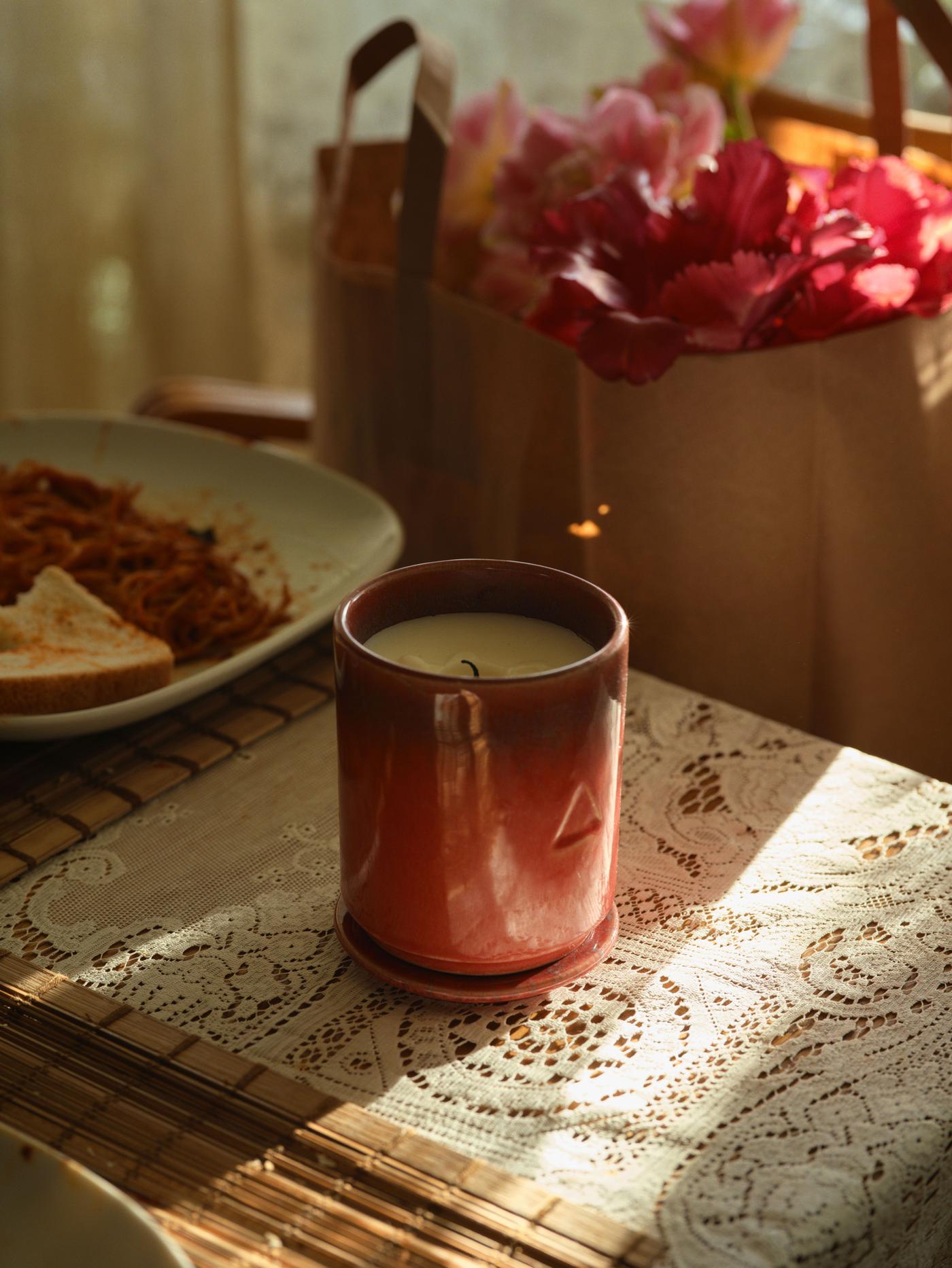 An unlit pink ceramic candle placed on a lace runner on a table. There are flowers in the background.