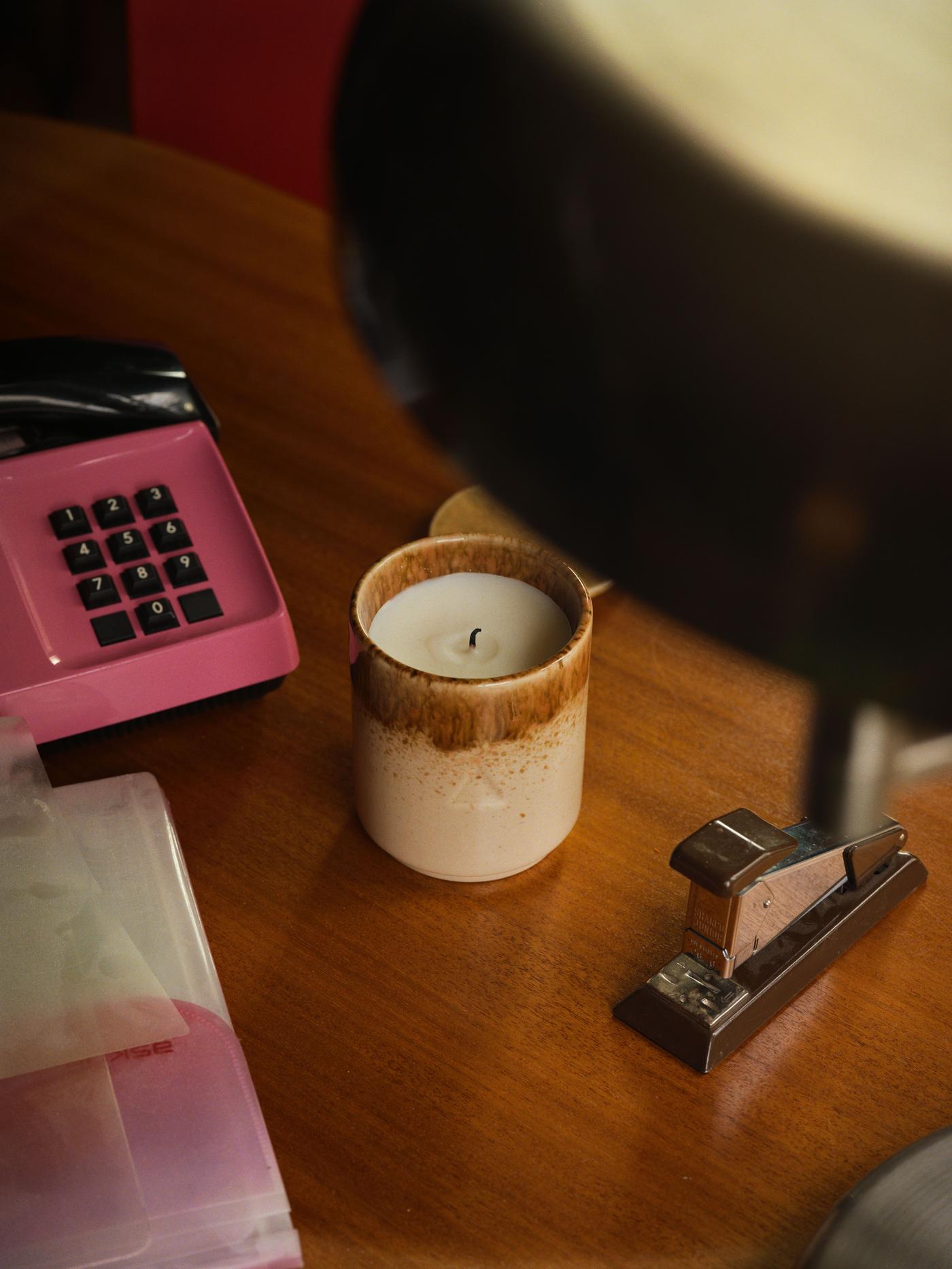 A brown and beige unlit candle on a wood table next to a stapler and retro telephone.