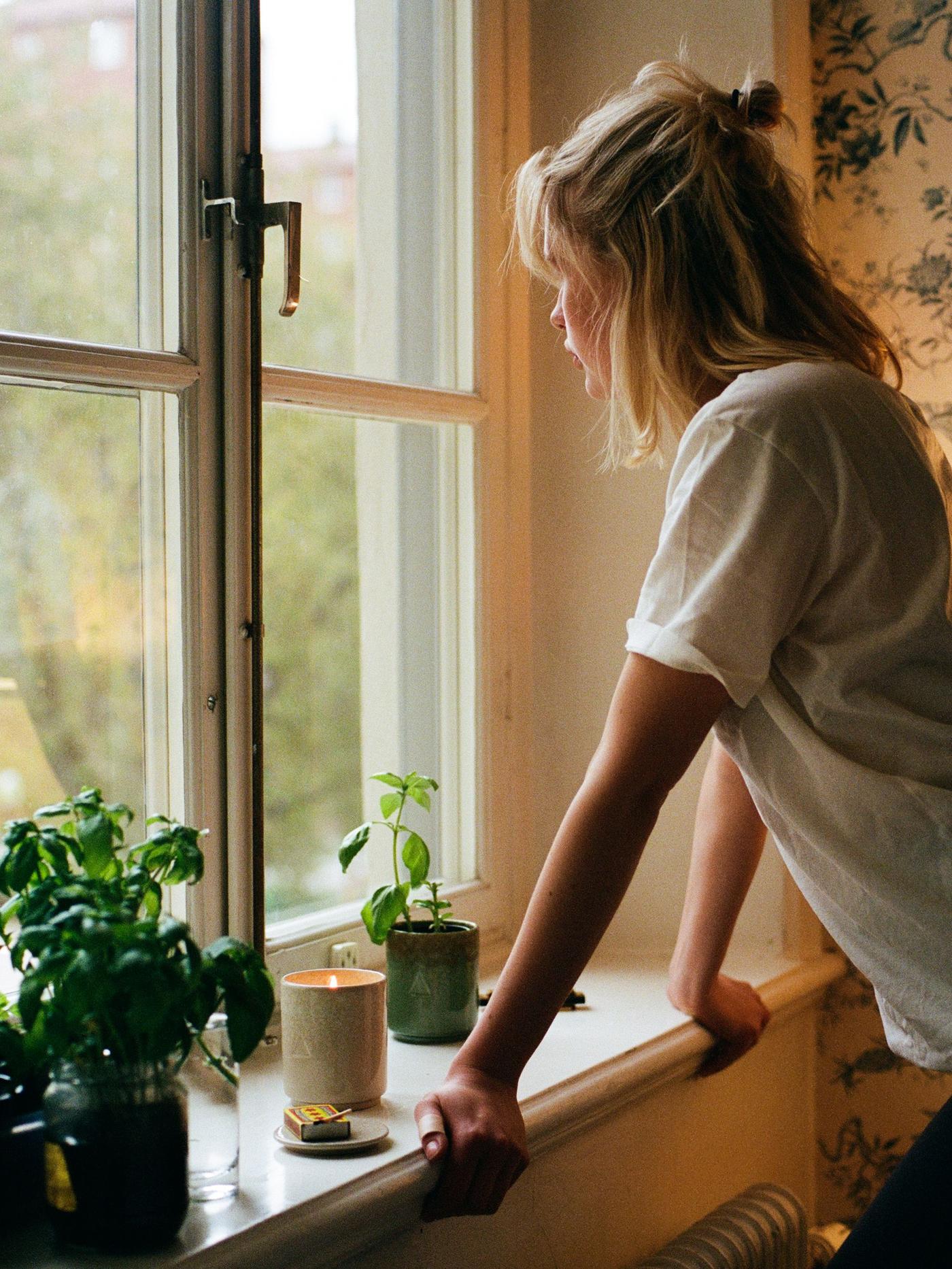 A person looks out of the window. There are plants and a burning candle on the windowsill.