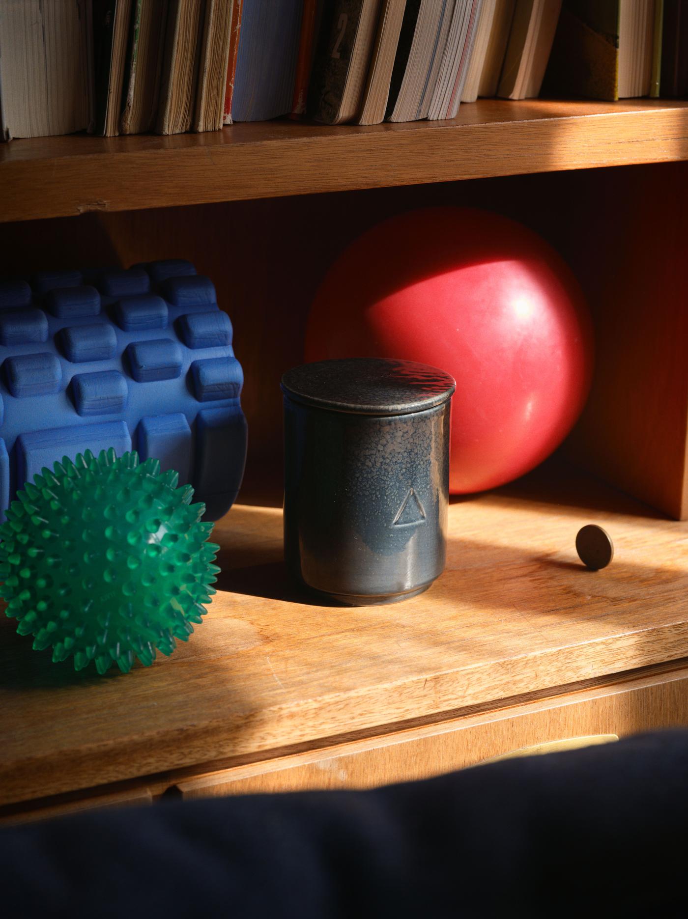 A dark blue ceramic candle with a lid sits on a wooden shelf alongside a green ball, a red ball and a blue object.