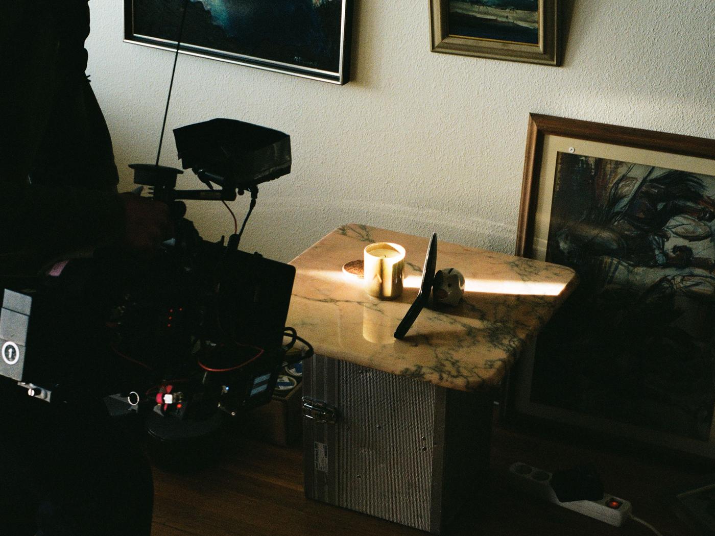 A professional video camera points at a beige candle on a marble table. There are framed pictures on the wall.