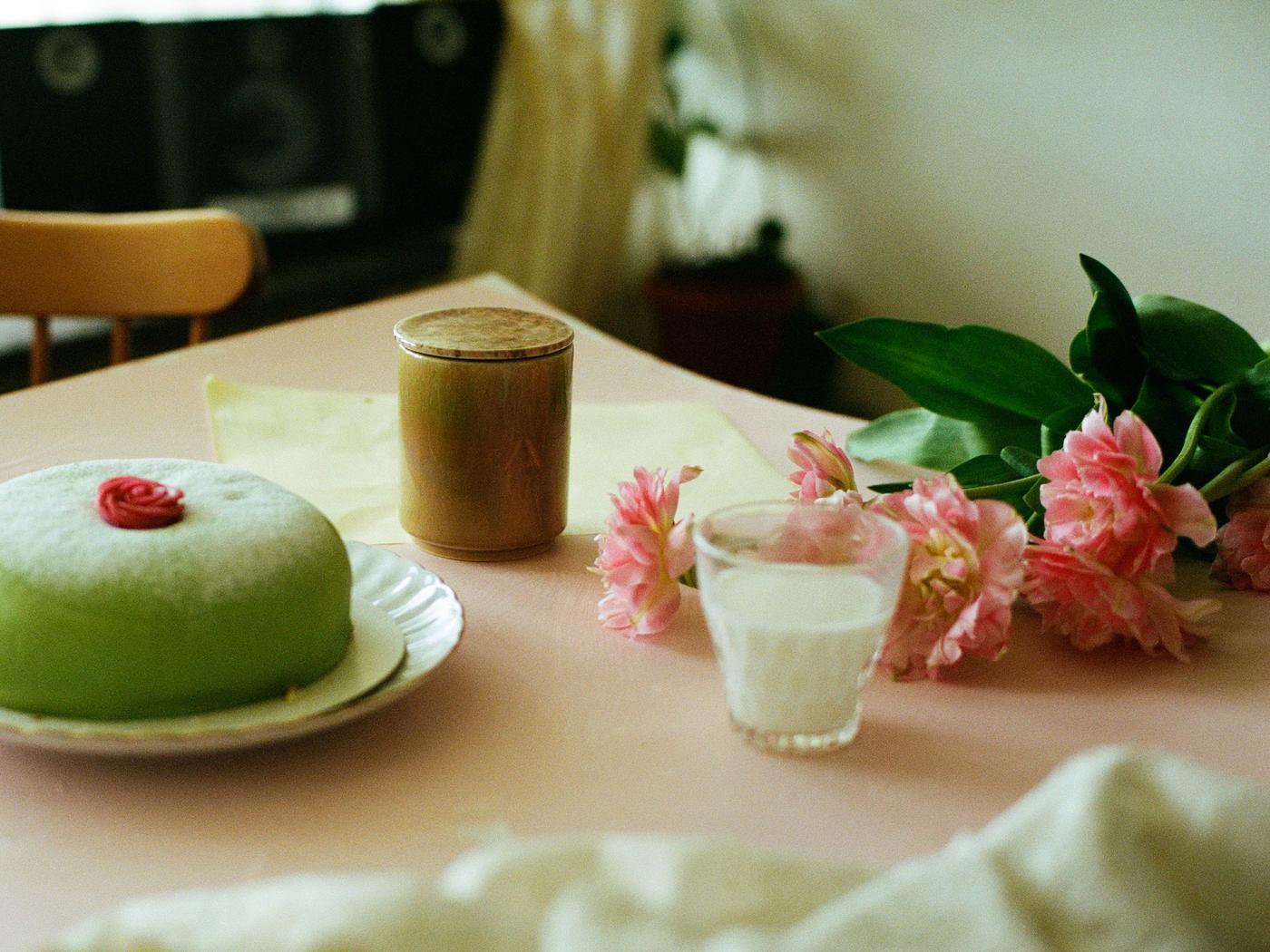 A table with a green princess cake, a glass of milk and flowers. A ceramic candle with lid sits next to them.