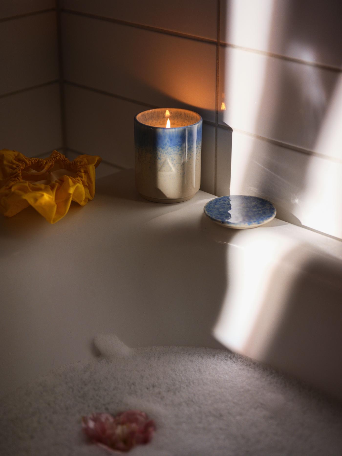 A blue and beige ceramic candle and tealights burn next to a bubble bath and a wood panel wall.