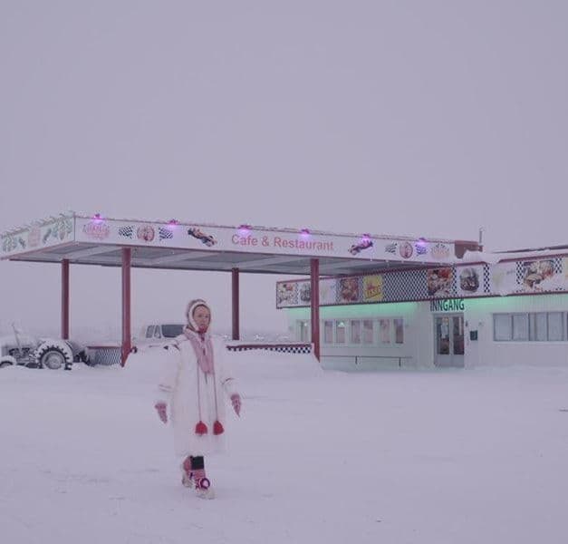 A person walks through the snow, past a roadside cafe and restaurant. The person is wearing winter clothes.