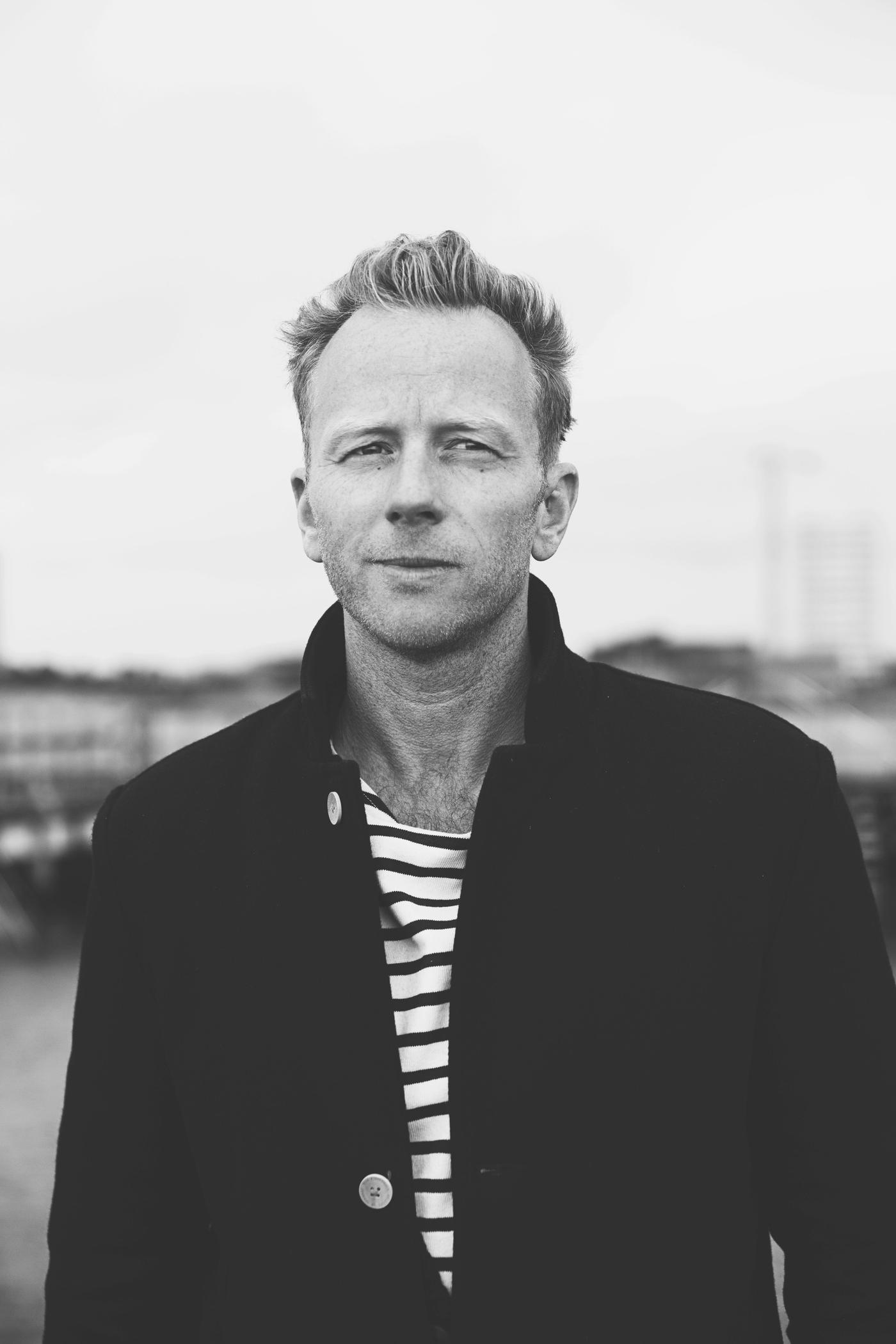 A black and white photo of a man standing outside. He wears a black jacket and striped top.