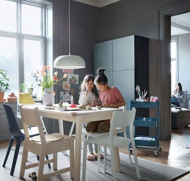 A child sits at a table, while another stands and looks over her. They are drawing in the dining area. A woman sits in the room next door, reading.