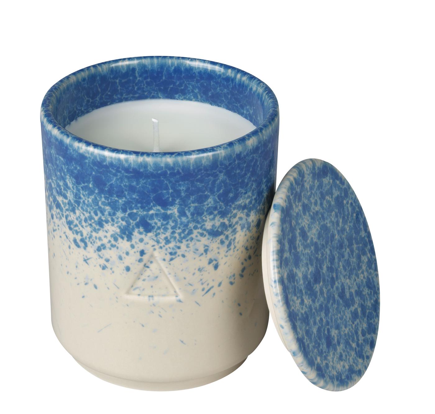 A beige and blue ceramic handle holder with a candle inside and a lid leaning against it. Against a white background.