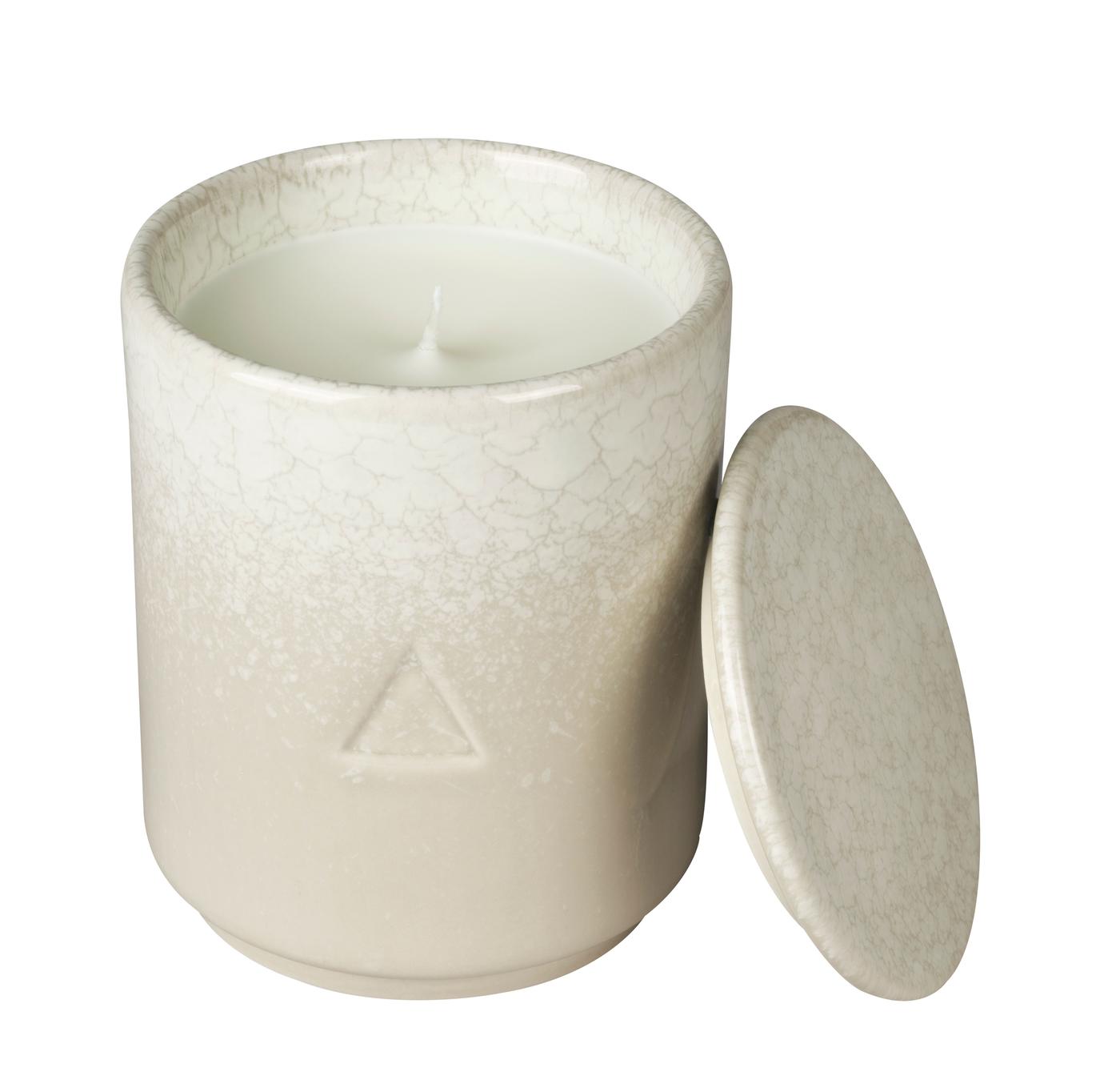 A cream and beige ceramic handle holder with a candle inside and a lid leaning against it. Against a white background.