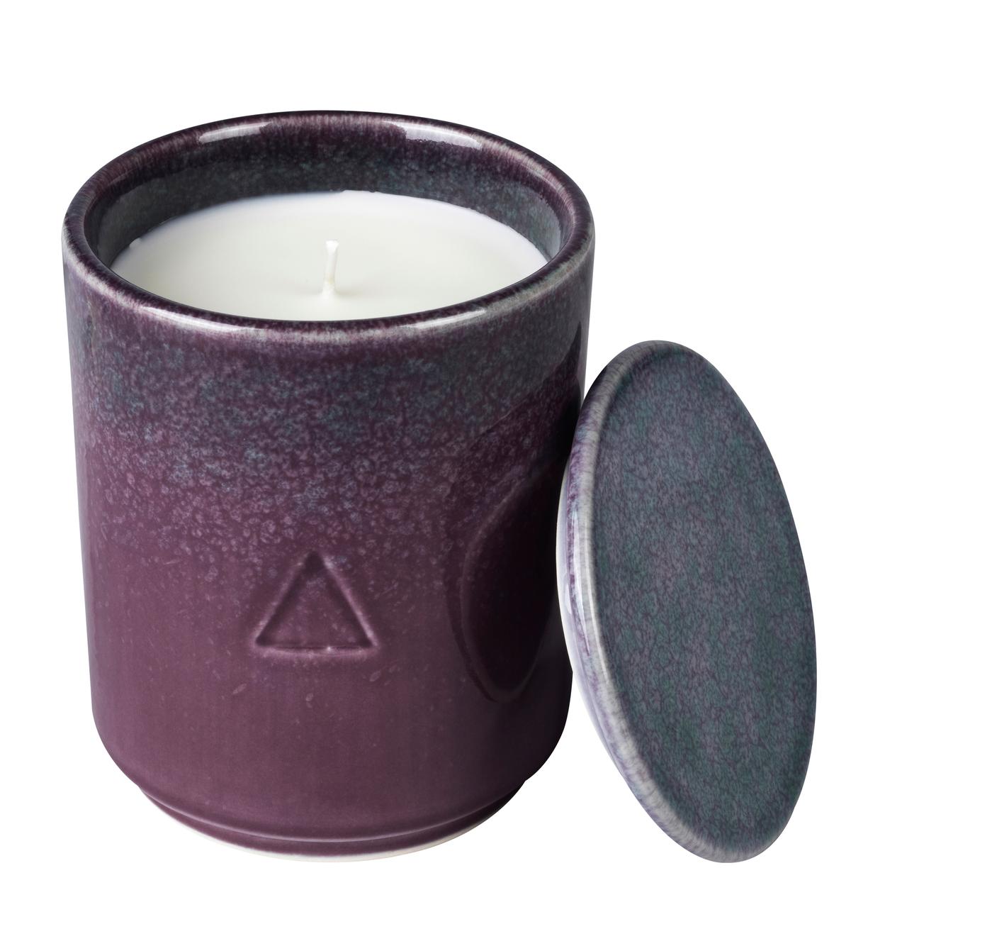 A dark brown and blue ceramic handle holder with a candle inside and a lid leaning against it. Against a white background.