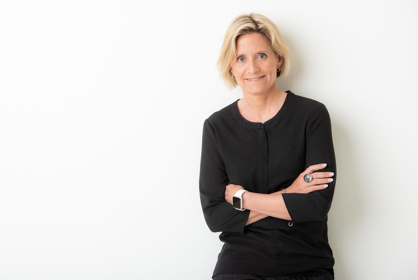 A woman in black clothes stands against a white background. Her arms are folded.