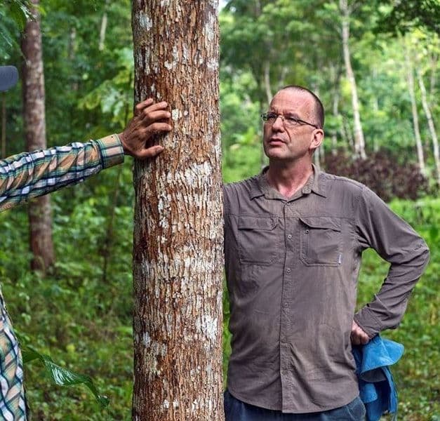 Two men look at a tree. One man with a cap has his hand on the tree.