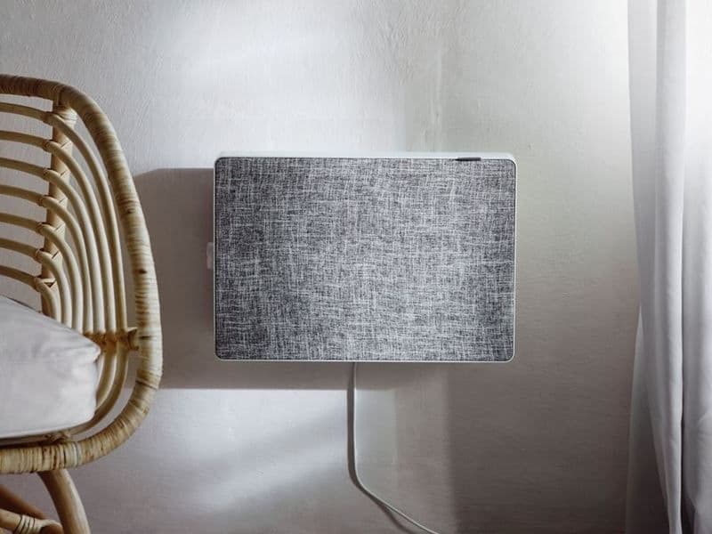 A grey air purifier is fixed to a pale wall. To the left is a chair and on the right is a white curtain.