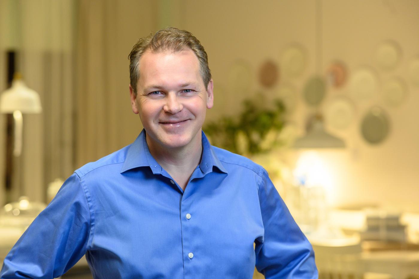 A man in a blue shirt smiles at the camera. He is in a room with lights on in the background.