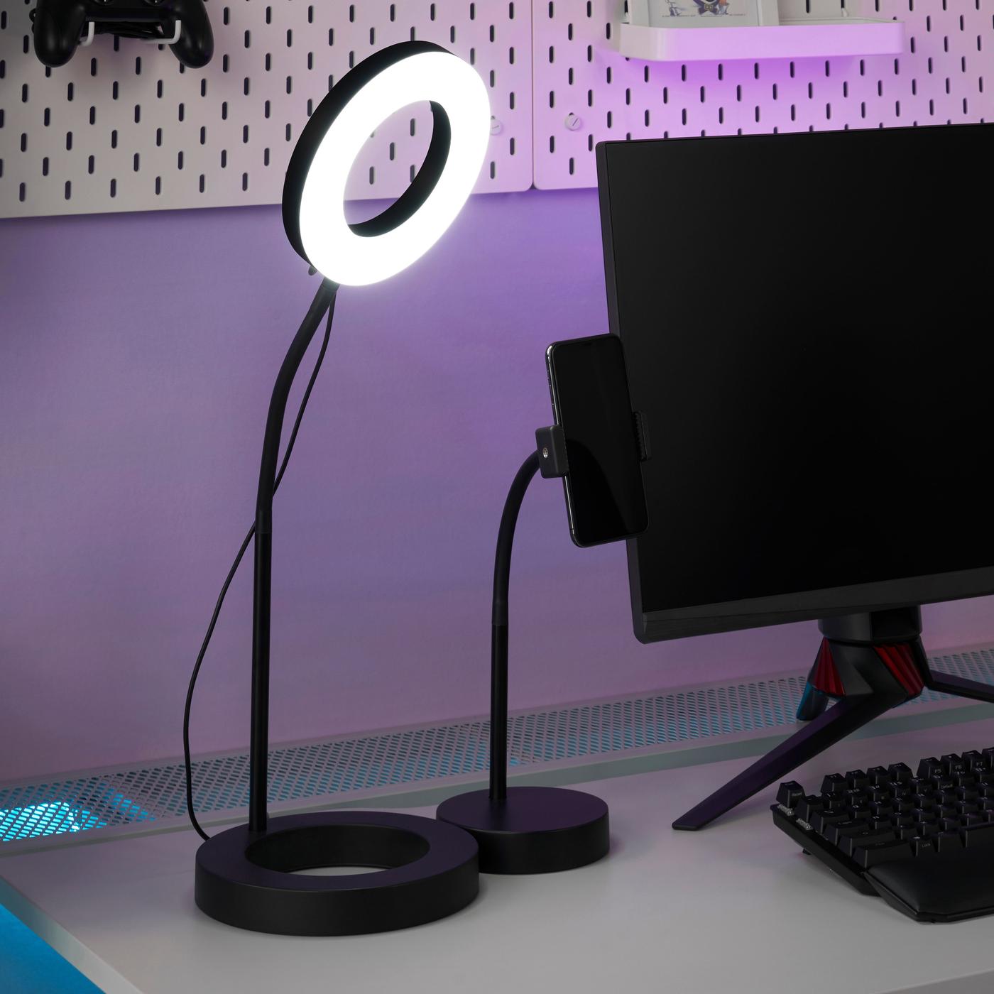 A black ring light is illuminated. It stands on a desk next to a black monitor.