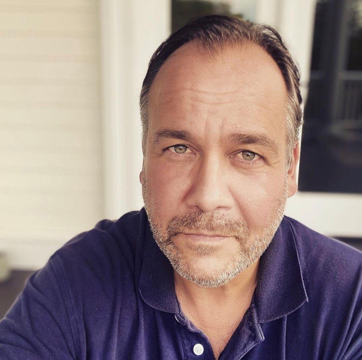 A man in a dark blue shirt looks at the camera. There is a window behind him.