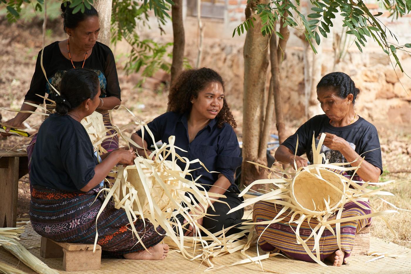 Four women sit on the floor outside, they are talking and weaving baskets out of palm leaves, surrounded by trees.