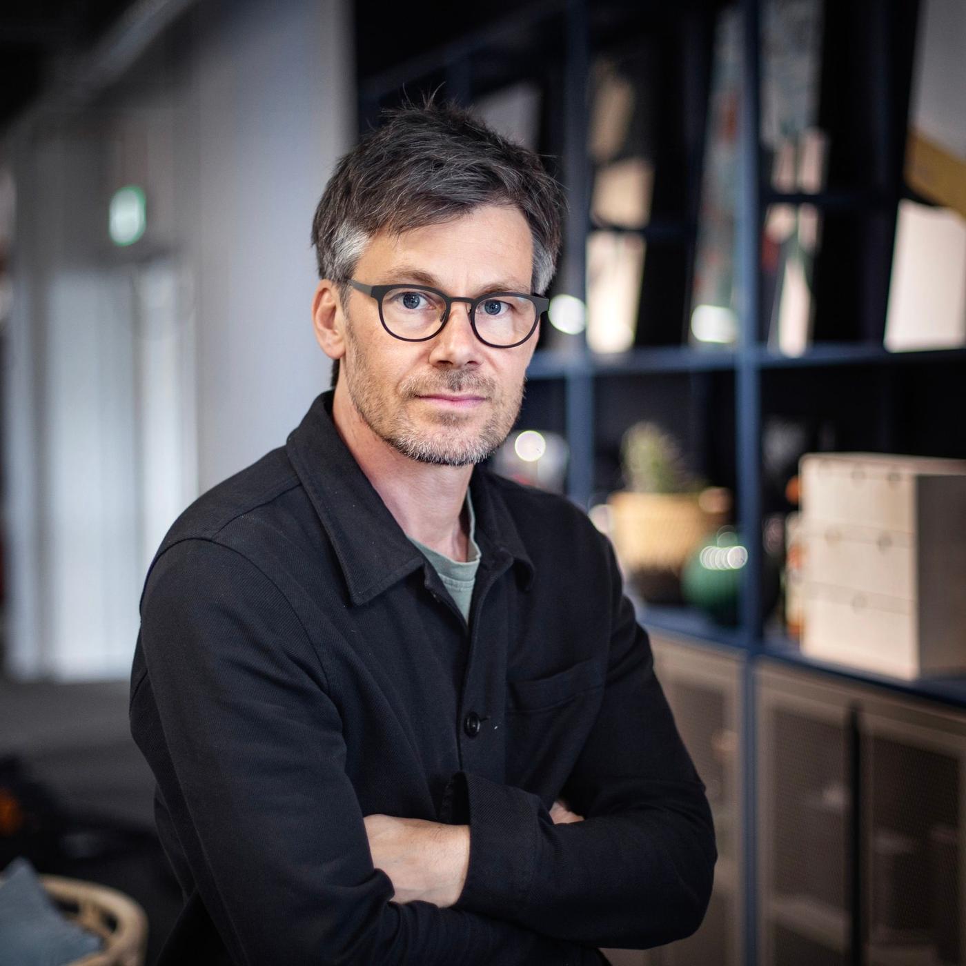 A man in a black shirt standing in a room with his arms crossed.