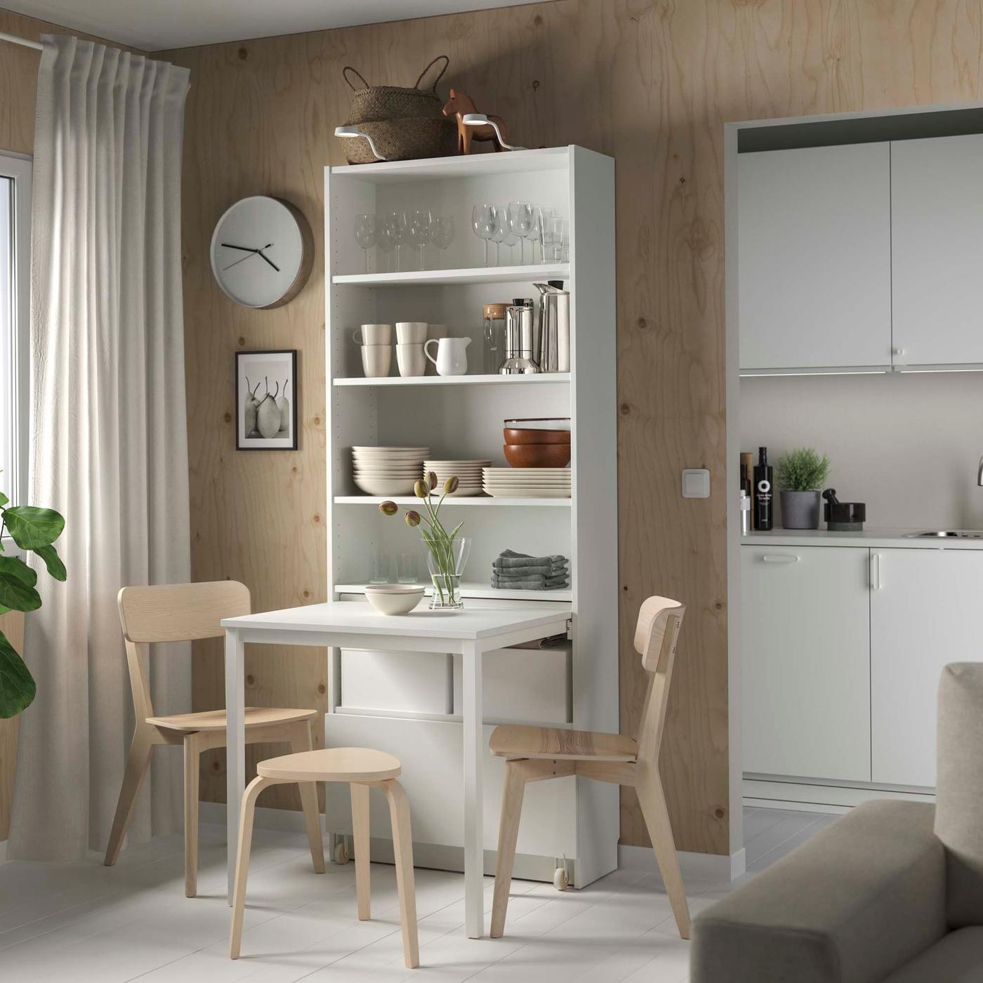 A small dining area with a white table set connected to a tall shelving unit holding various kitchenware.
