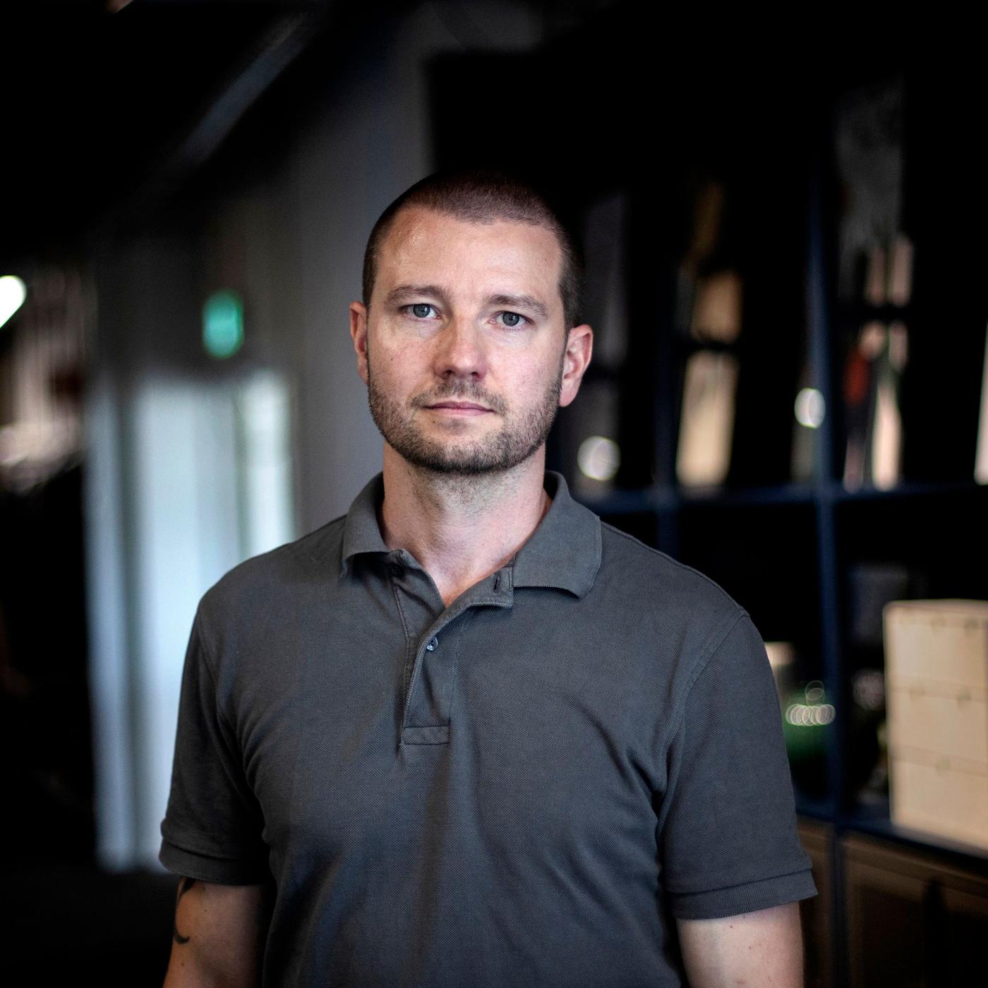 A portrait of a man in a grey shirt standing in an office environment.