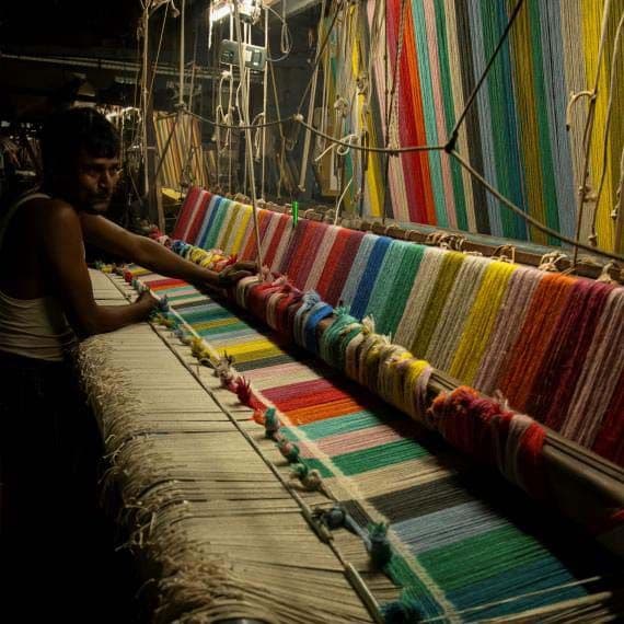 A person stands at a long loom, weaving a brightly coloured striped rug.
