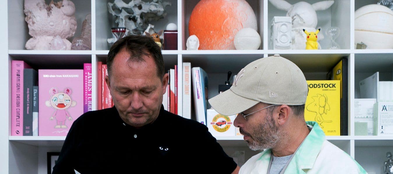 Two men looking at a piece of paper at a desk with decoration, in front of a white shelving unit with books and decorations.