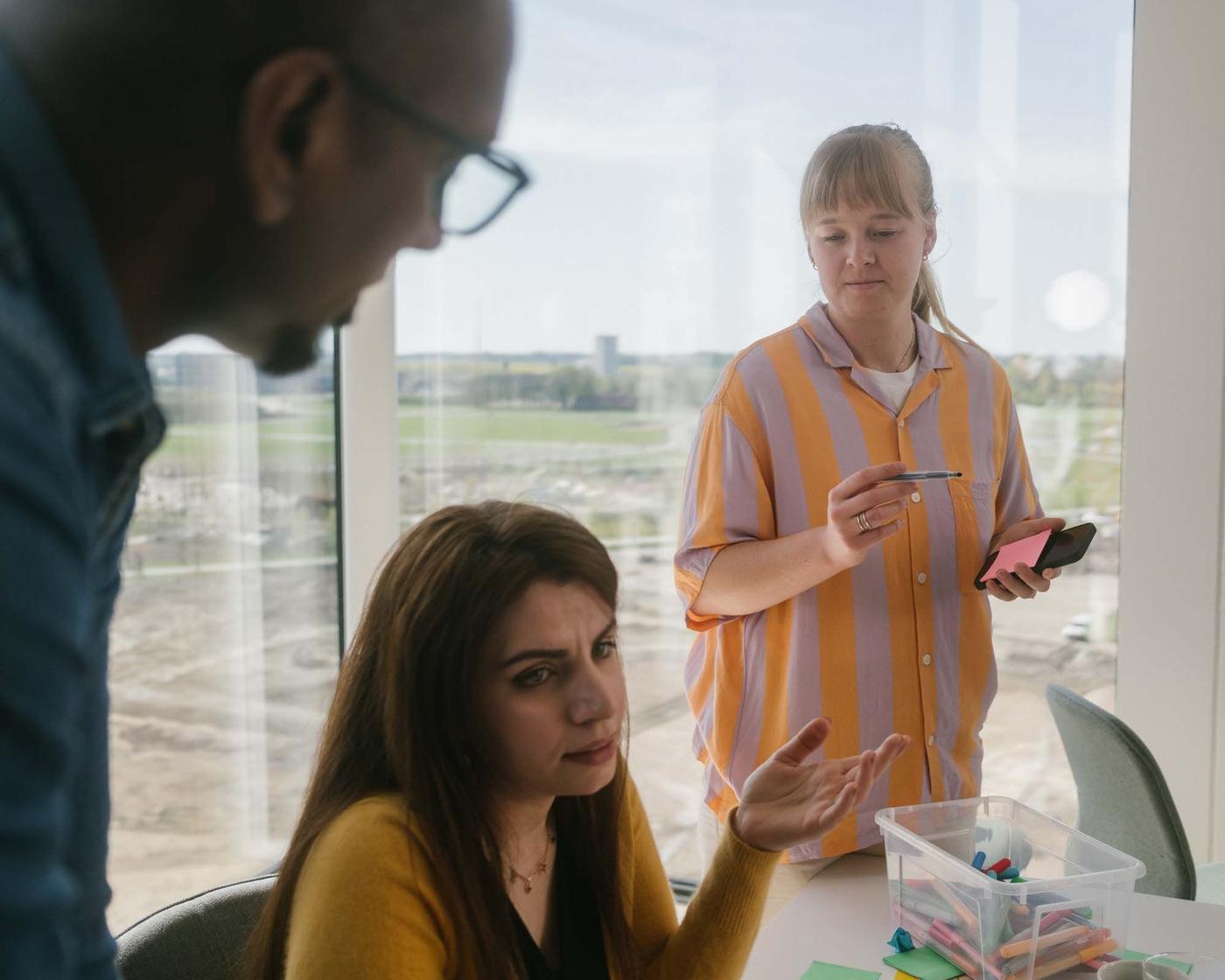A woman stands, another woman sits, while a man in the foreground looks at the woman as she speaks.