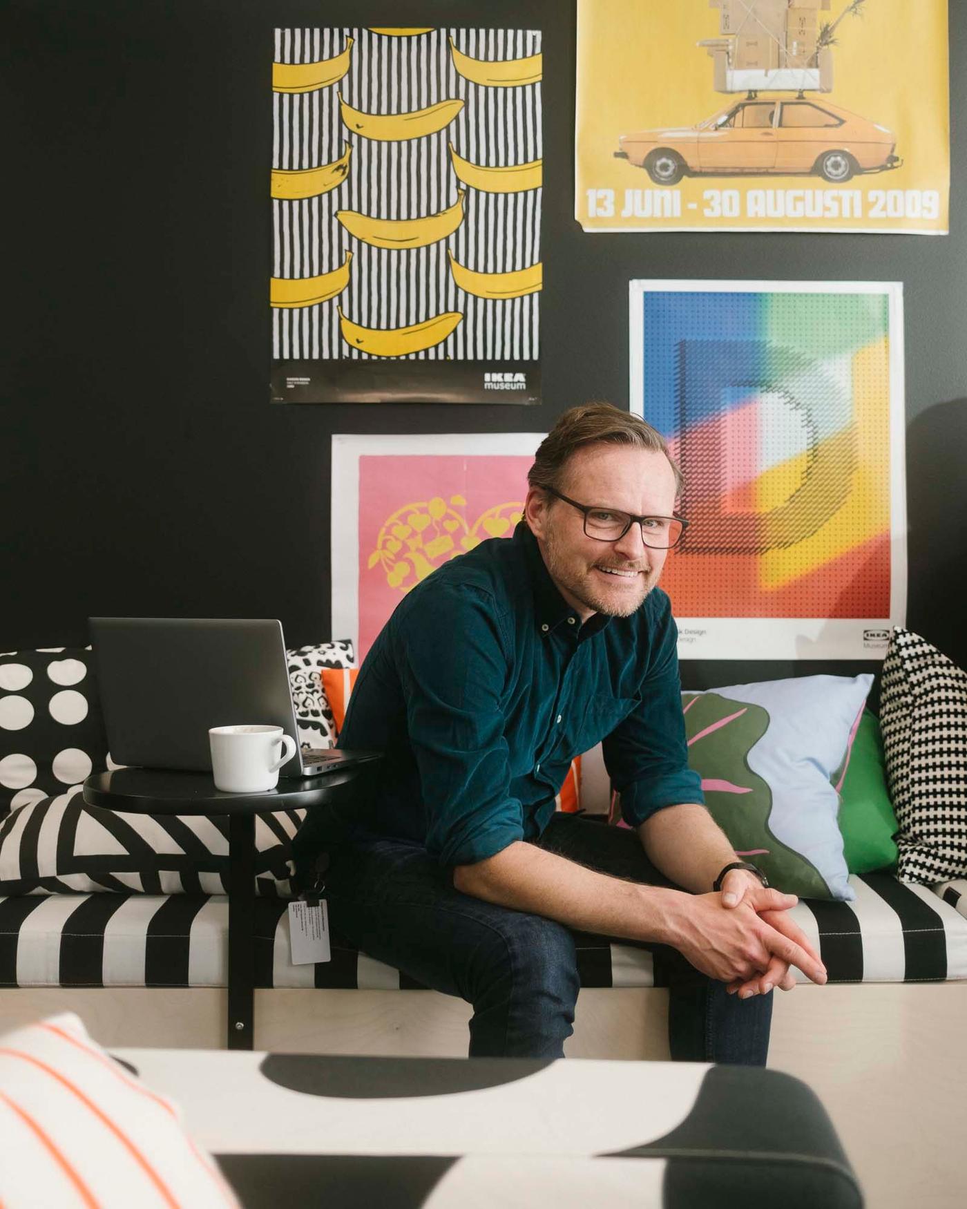 A man sits on a striped seat with pictures on the wall. He is smiling.