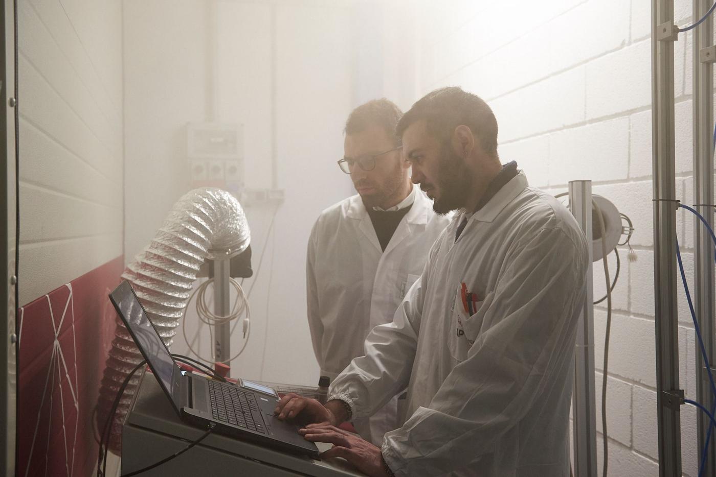 Two men in white coats working in front of a laptop computer at a testing facility.
