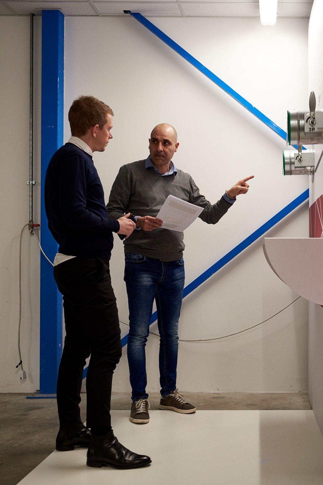 Two men engaged in conversation in a white room.