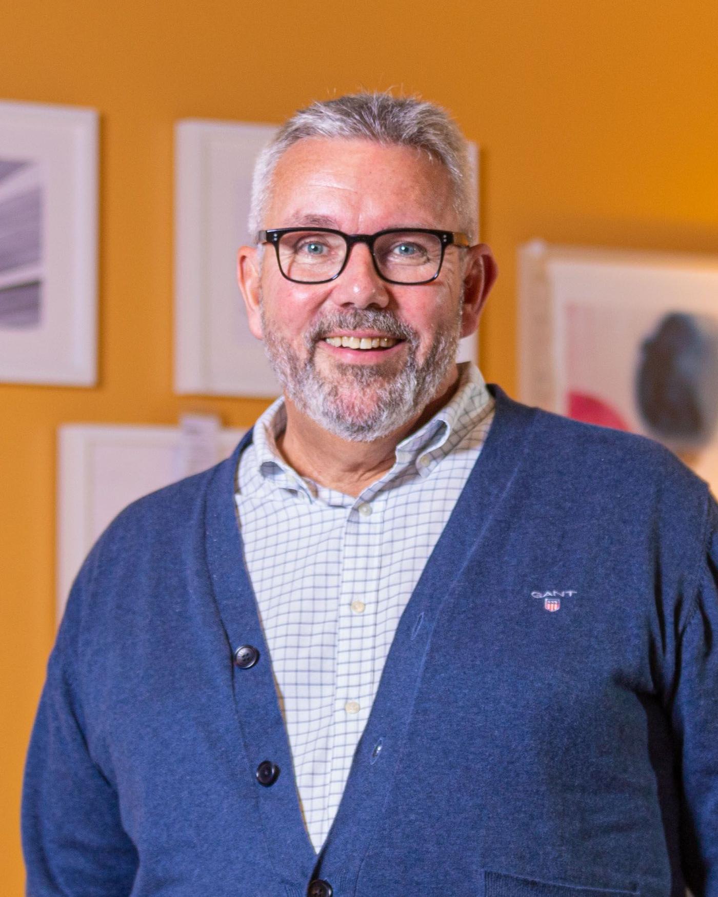 Portrait of Dennis Balslev standing against an orange wall filled with white picture frames.