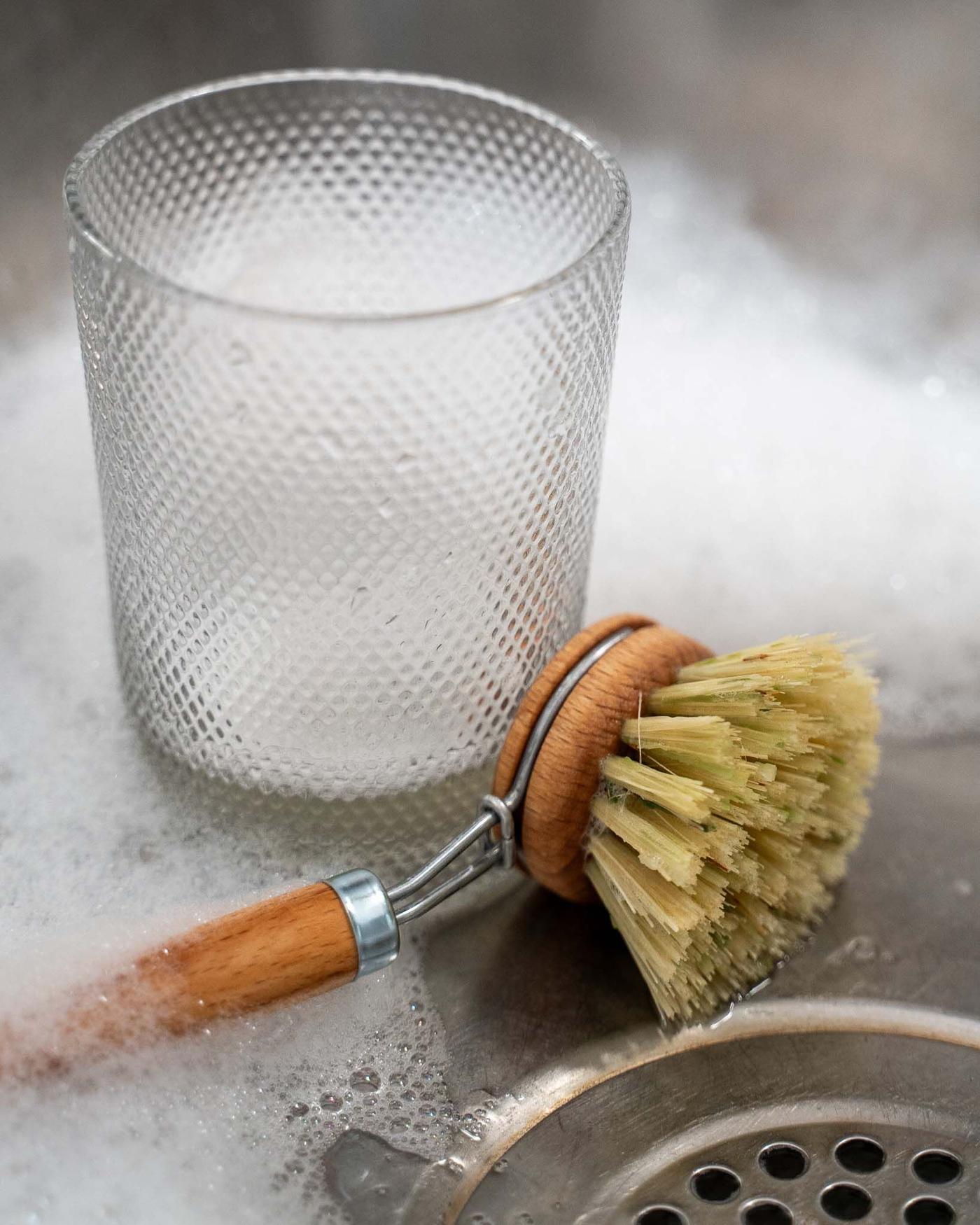 A glass and dish brush in a stainless steel sink surrounded by soap suds.