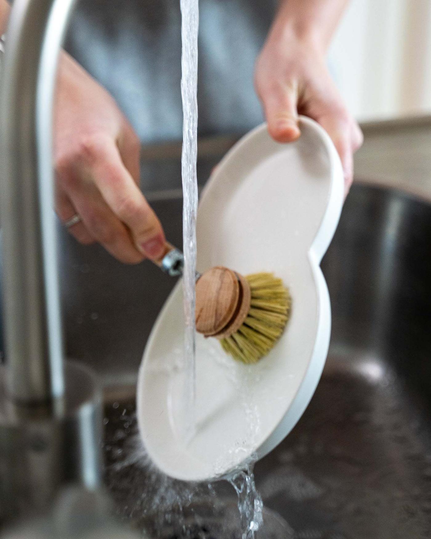 A close-up of a person doing the dishes with a brush under a stainless steel tap.