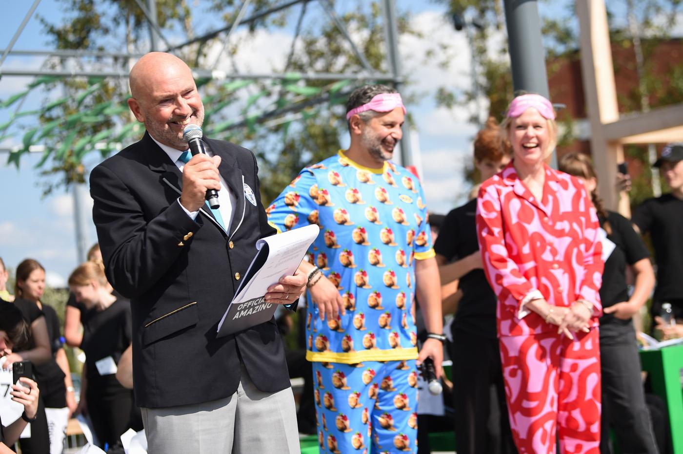 A man in suit on the mic next to a man in blue pyjama and a woman in red pyjama, on stage