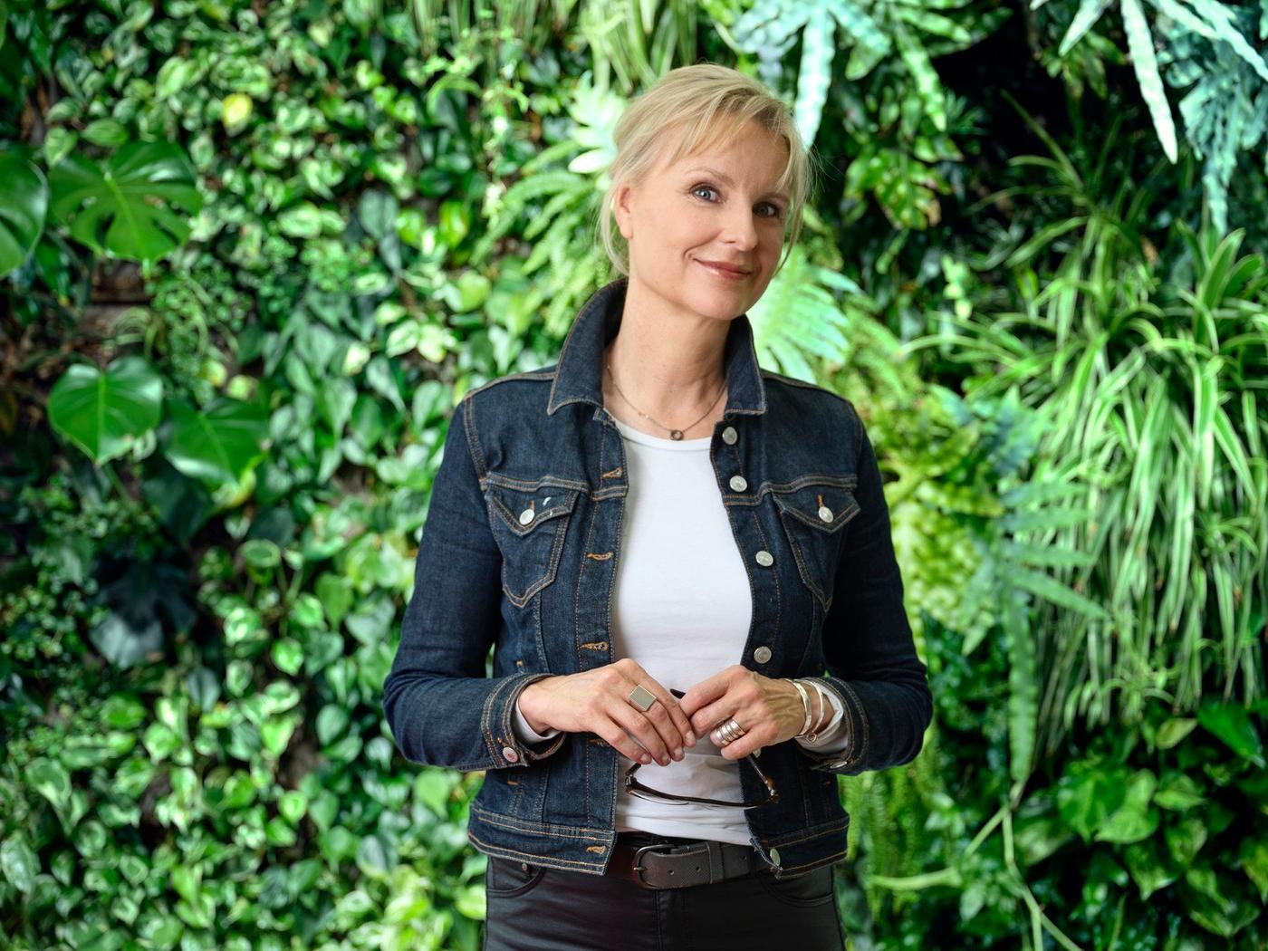 A woman in a white T-shirt and jeans jacket standing in front of a wall of green plants.