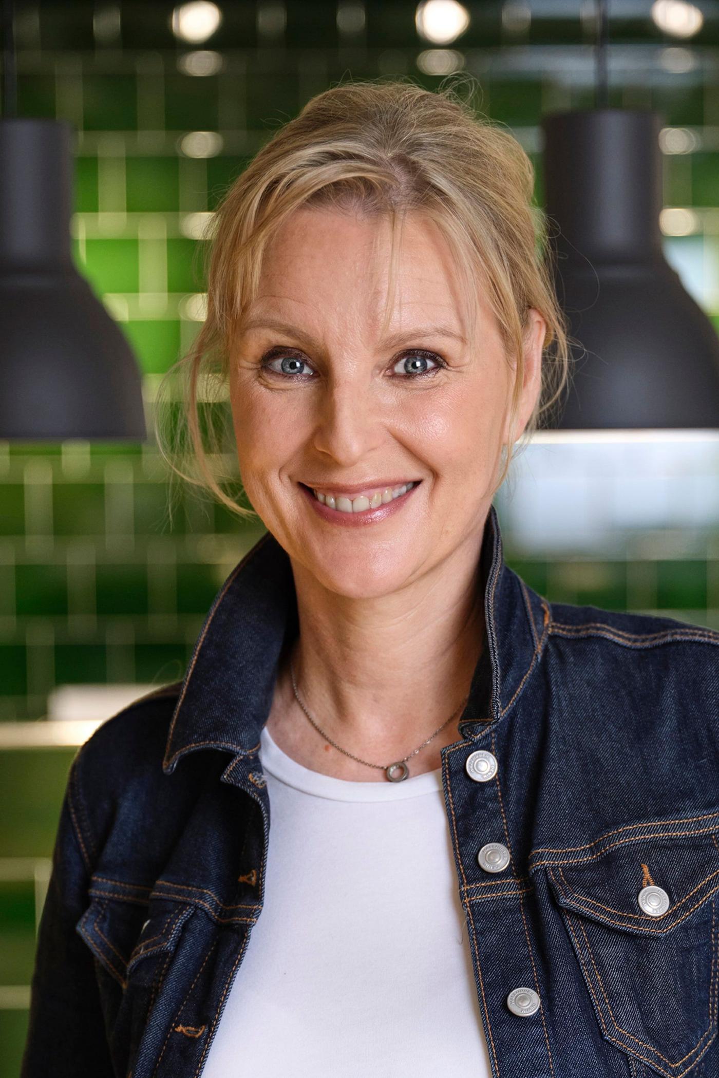 A woman in a white T-shirt and jeans jacket standing in a room. In the background are green tiles.